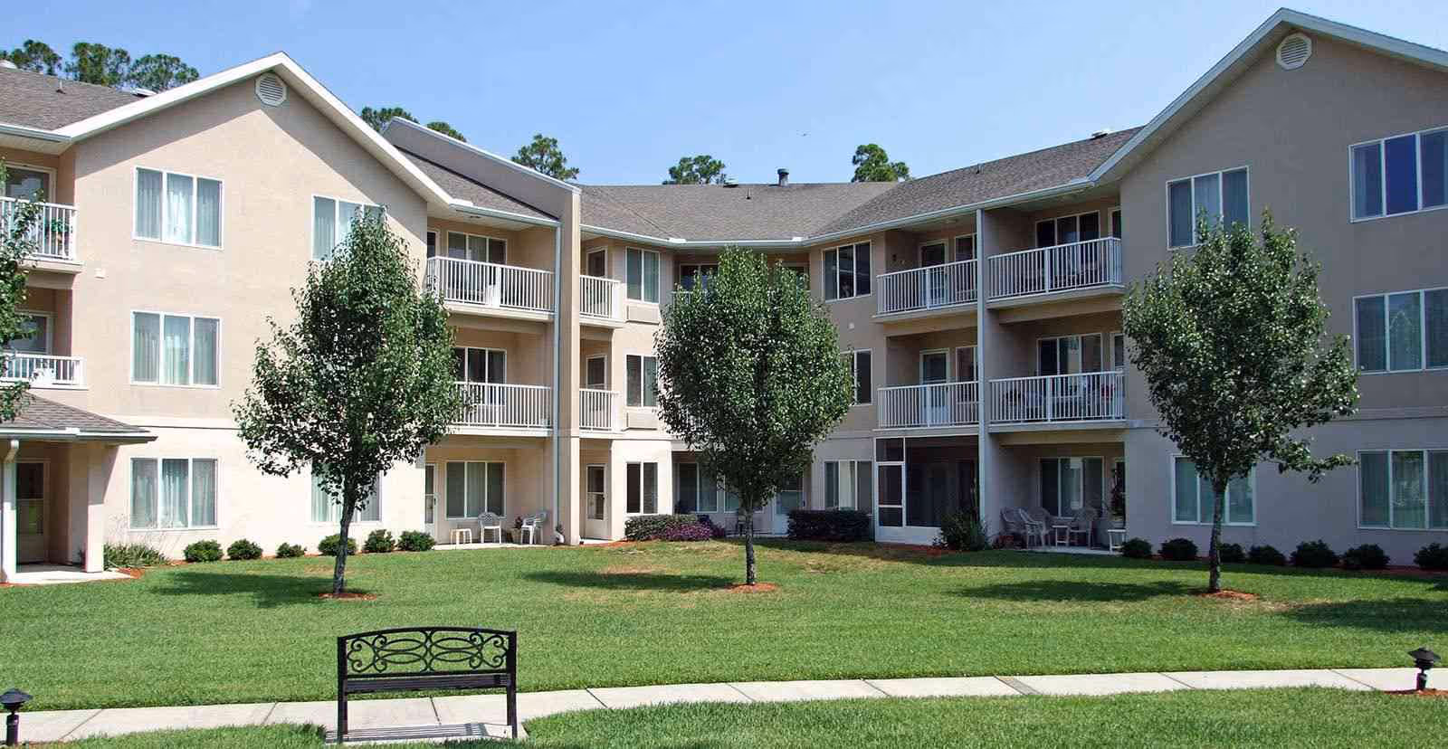 Exterior view of a three-story senior living facility building with beige walls and white railings on balconies. There is a well-maintained green lawn with three small trees and a black metal bench in the foreground under a clear blue sky.