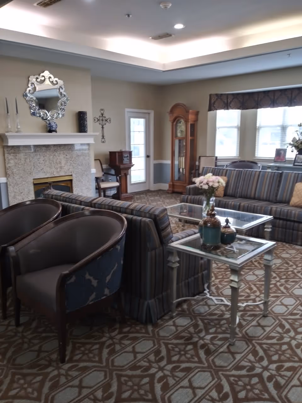 A cozy living room with patterned carpet, two striped sofas, two armchairs, and two glass-top tables with decorative vases and flowers. There is a fireplace with a decorative mirror above it, a grandfather clock, and a door with glass panels. The room has large windows with valances and soft lighting from a recessed ceiling.