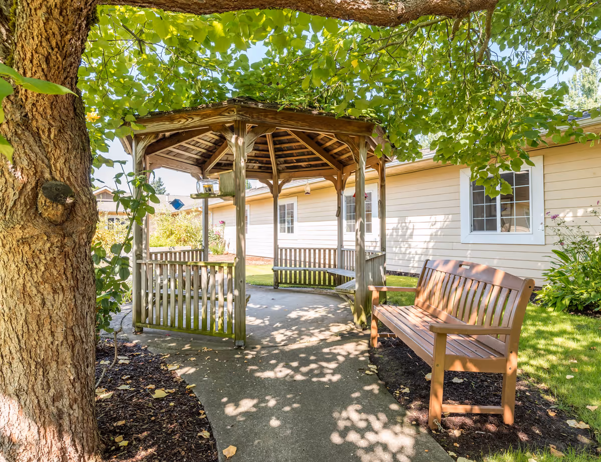 A wooden gazebo with benches inside and a wooden bench outside, situated in a garden area with green grass, plants, and a large tree providing shade. The exterior of a light-colored building with white-framed windows is visible in the background.
