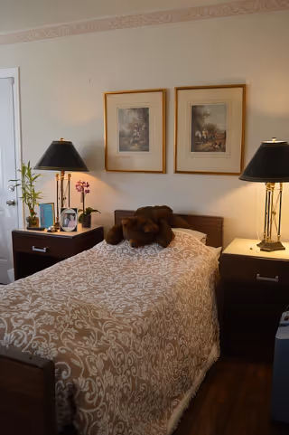 A cozy bedroom with a single bed covered in a beige and white patterned bedspread. A brown teddy bear is placed on the bed. On each side of the bed are dark wooden nightstands with black lamps. The left nightstand also has a small plant, a framed photo, and a book. Two framed paintings hang on the wall above the bed. The room has light-colored walls and wooden flooring.