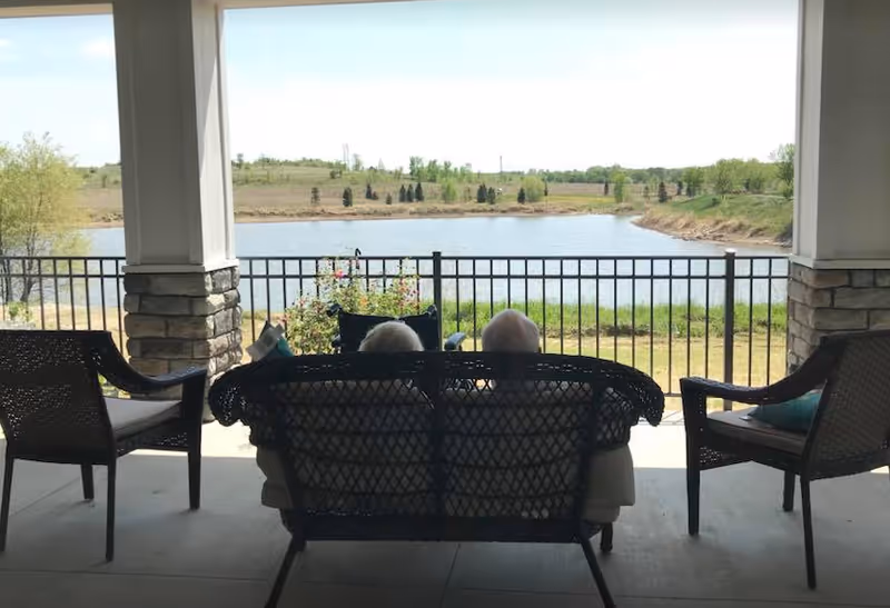 Two people sitting on a covered patio bench facing a lake and landscaped grounds.