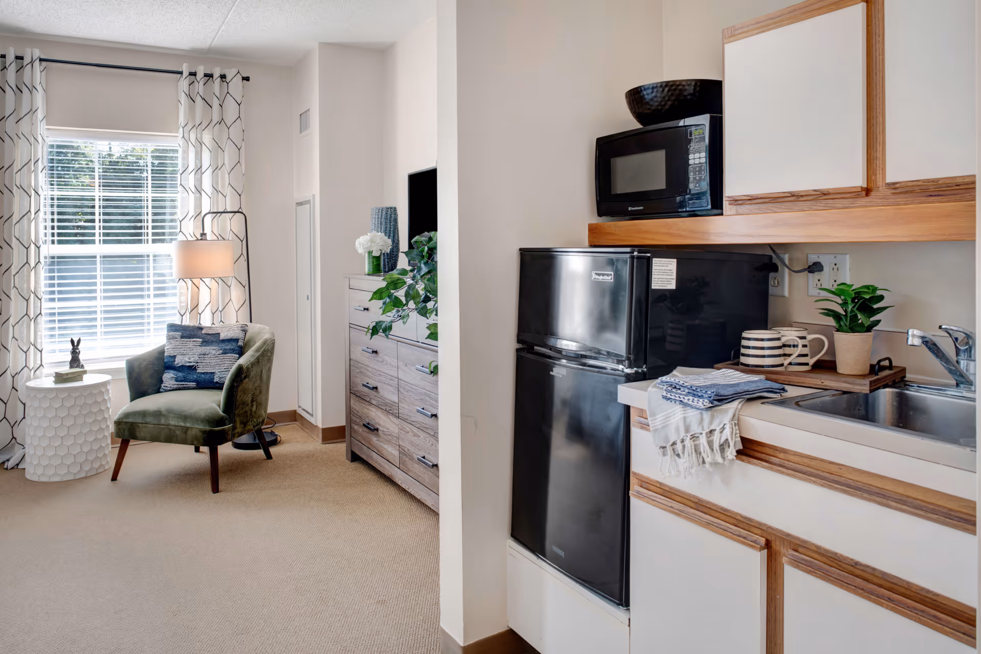 A cozy interior space featuring a small kitchenette with a black mini refrigerator, microwave, sink, and wooden cabinets. To the left, there is a sitting area with a green upholstered chair, a small white side table with a decorative item, a floor lamp, and a dresser with plants and decorative vases. A window with patterned curtains allows natural light to brighten the room.