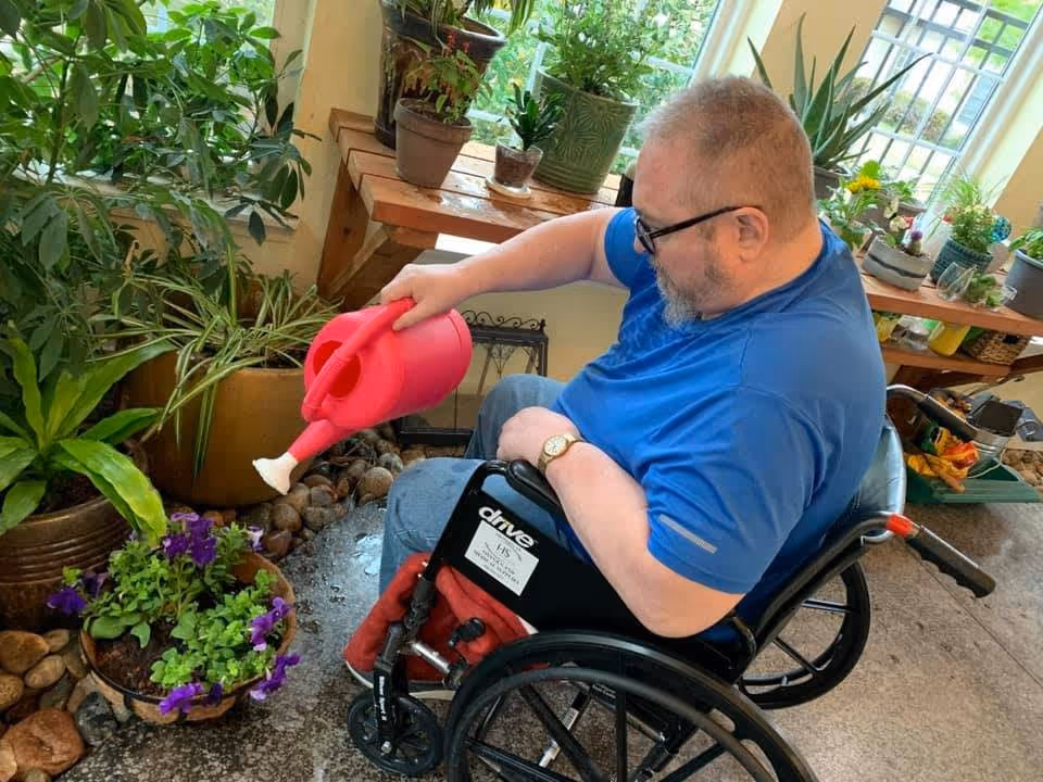 A man in a wheelchair wearing a blue shirt waters plants indoors using a red watering can. The area is filled with various potted plants and natural light coming through the windows.
