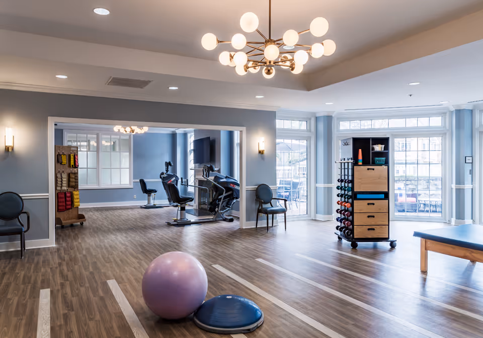 A spacious and well-lit exercise room in a senior living facility with wooden flooring and light blue walls. The room contains exercise equipment including a recumbent bike, a rack with various dumbbells, a purple exercise ball, and a blue balance trainer. There are several chairs along the walls and large windows letting in natural light. A modern chandelier hangs from the ceiling.