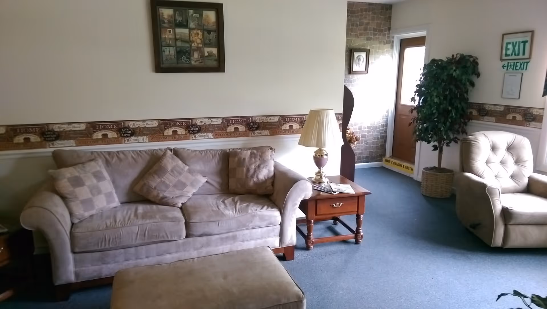 A cozy living room with a beige sofa adorned with three checkered cushions, a matching ottoman, and a beige armchair. A wooden side table with a lamp and some magazines sits next to the sofa. The walls are decorated with a framed picture and a wallpaper border featuring home-themed words. There is a potted plant near a door with exit signs above it, and the floor is carpeted in blue.