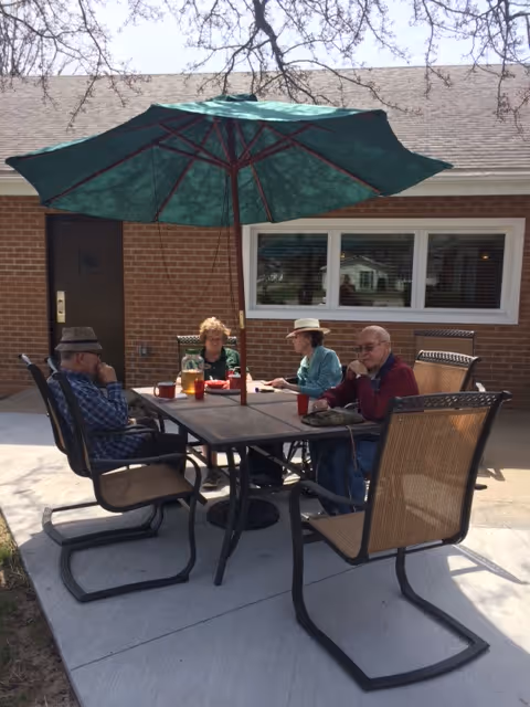 Four elderly people sitting around a rectangular outdoor table under a large green patio umbrella. They are seated on cushioned chairs on a concrete patio in front of a brick building with a window and door visible in the background. The group appears to be engaged in conversation or an activity together.