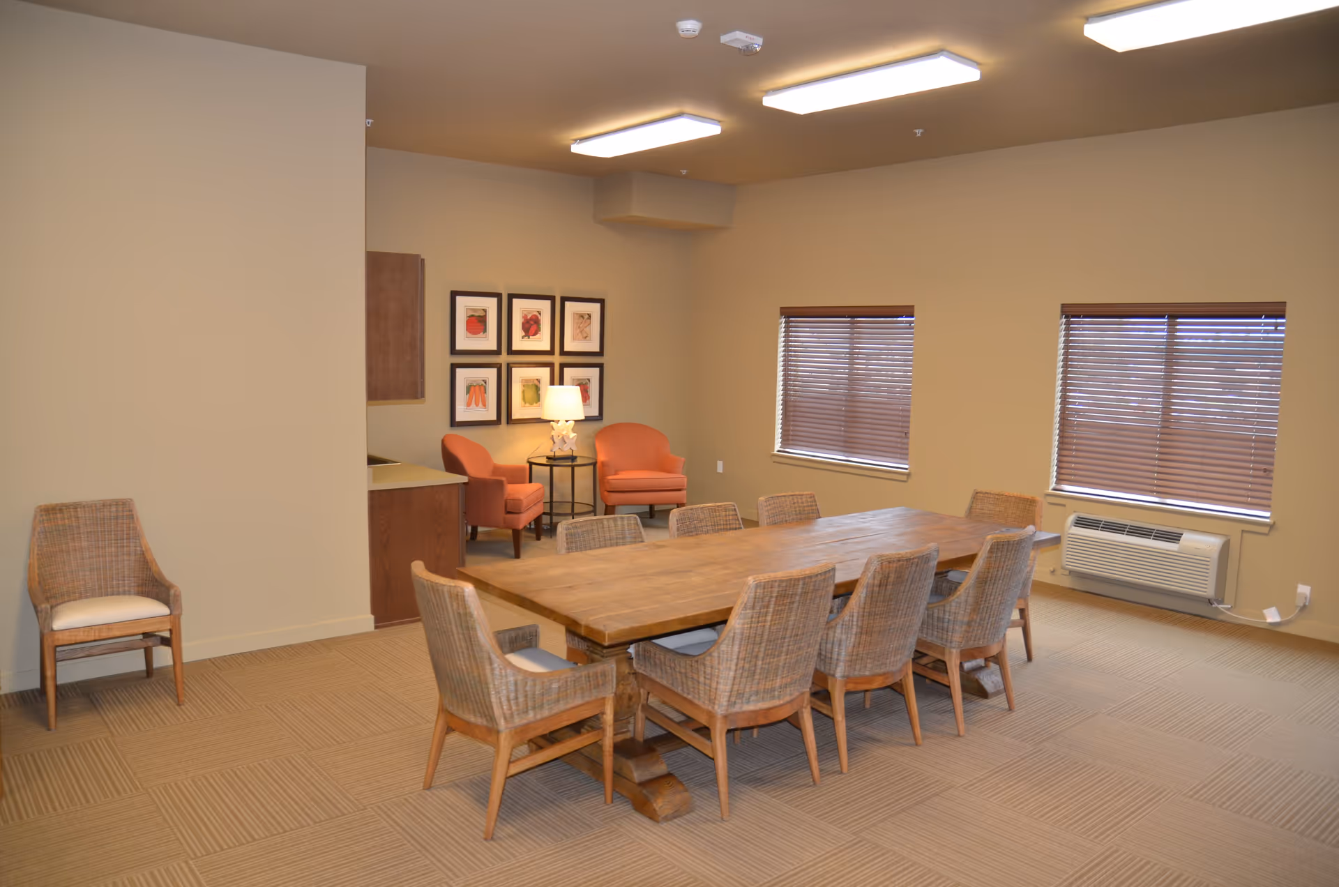 A well-lit room with a large wooden table surrounded by eight wicker chairs with cushions. In the corner, there are two orange upholstered armchairs with a small round table and a lamp between them. The walls are beige with six framed pictures hanging above the armchairs. Two windows with closed blinds and a wall-mounted air conditioning unit are visible on the right side. The floor is carpeted with a striped pattern.