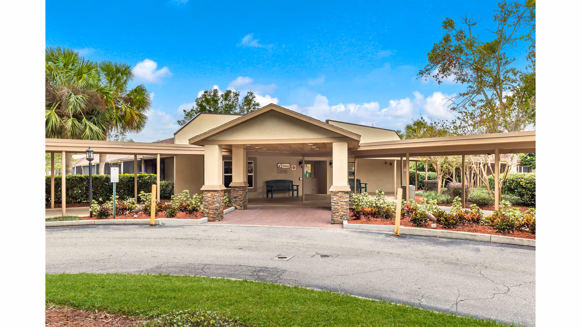 Exterior view of a single-story building entrance with a covered drop-off area supported by stone pillars. The building is surrounded by landscaped bushes, trees, and a circular driveway under a bright blue sky with some clouds.