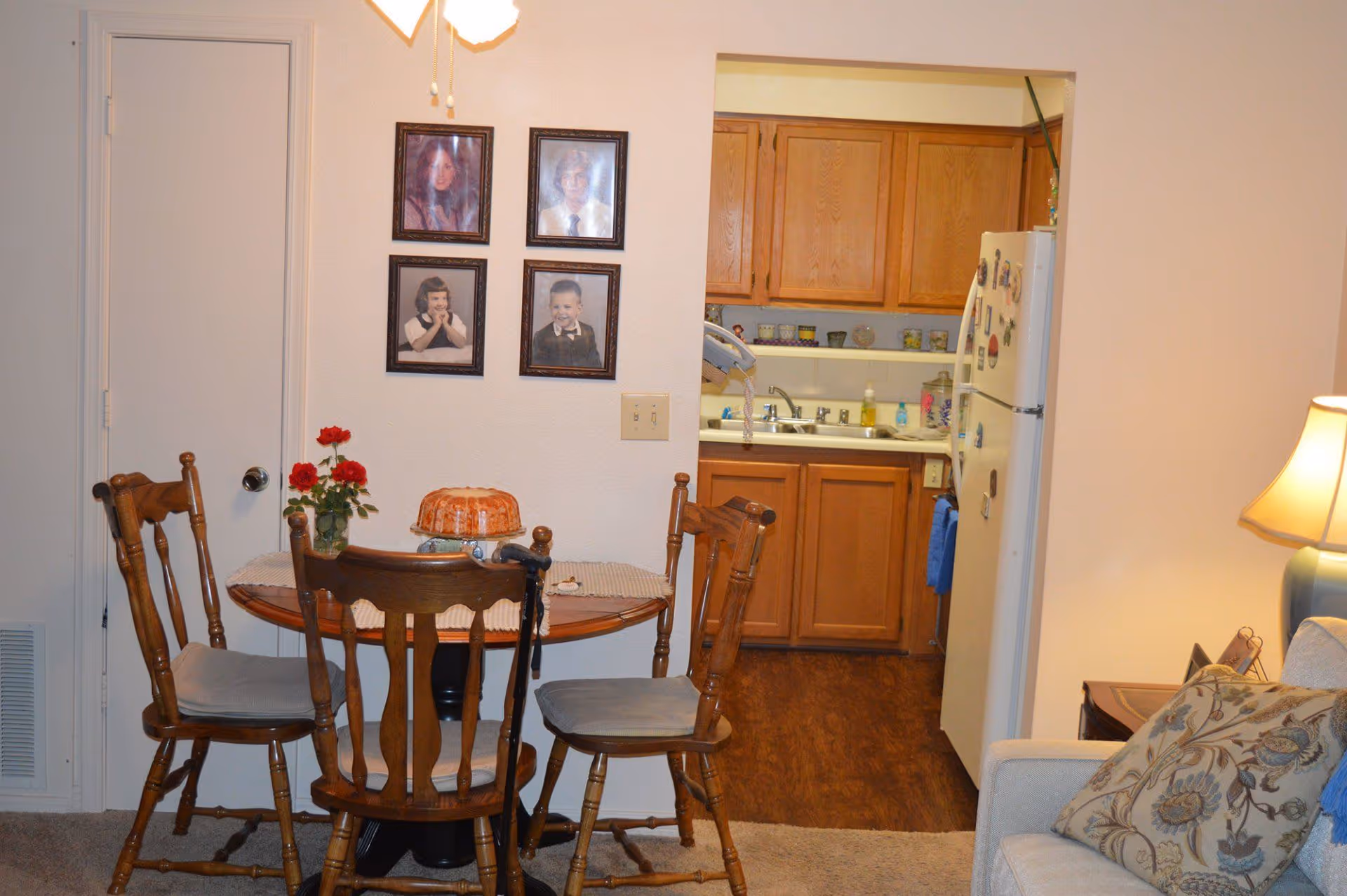 A cozy dining area with a round wooden table and four wooden chairs with cushions. On the table, there is a vase with red flowers and a bundt cake. Behind the table, there are four framed portraits on the wall. To the right, there is a view into a kitchen with wooden cabinets, a white refrigerator with magnets, a sink, and various kitchen items. A beige couch with patterned pillows and a lit table lamp are partially visible on the right side.