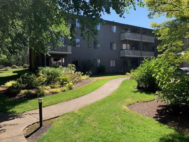 A three-story residential building with balconies surrounded by green lawns, trees, and landscaped bushes under a clear blue sky.