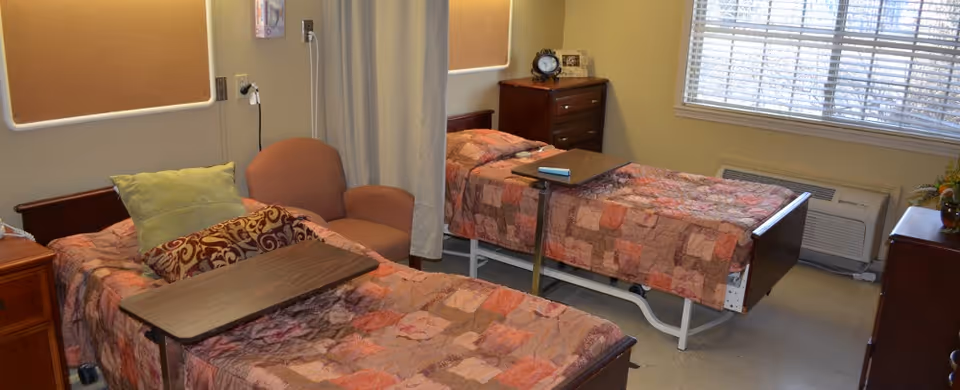 A room with two single beds covered with patterned quilts, each with an overbed table. There is a cushioned armchair between the beds, a wooden dresser with a clock and picture frame, and a window with blinds letting in natural light. The room has beige walls and a curtain that can be drawn for privacy.