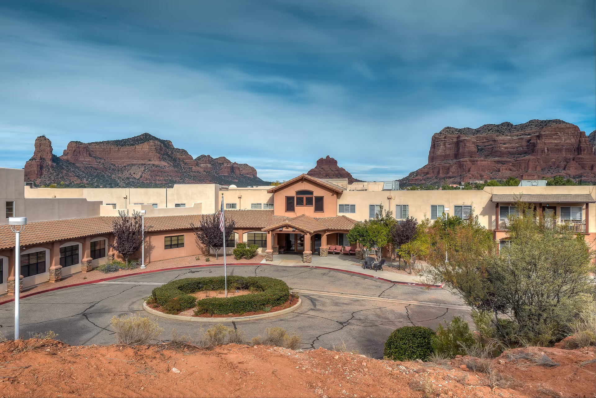 Front exterior view of Sedona Winds Assisted Living Community with a circular driveway and landscaped bushes, set against a backdrop of red rock formations and a partly cloudy sky.