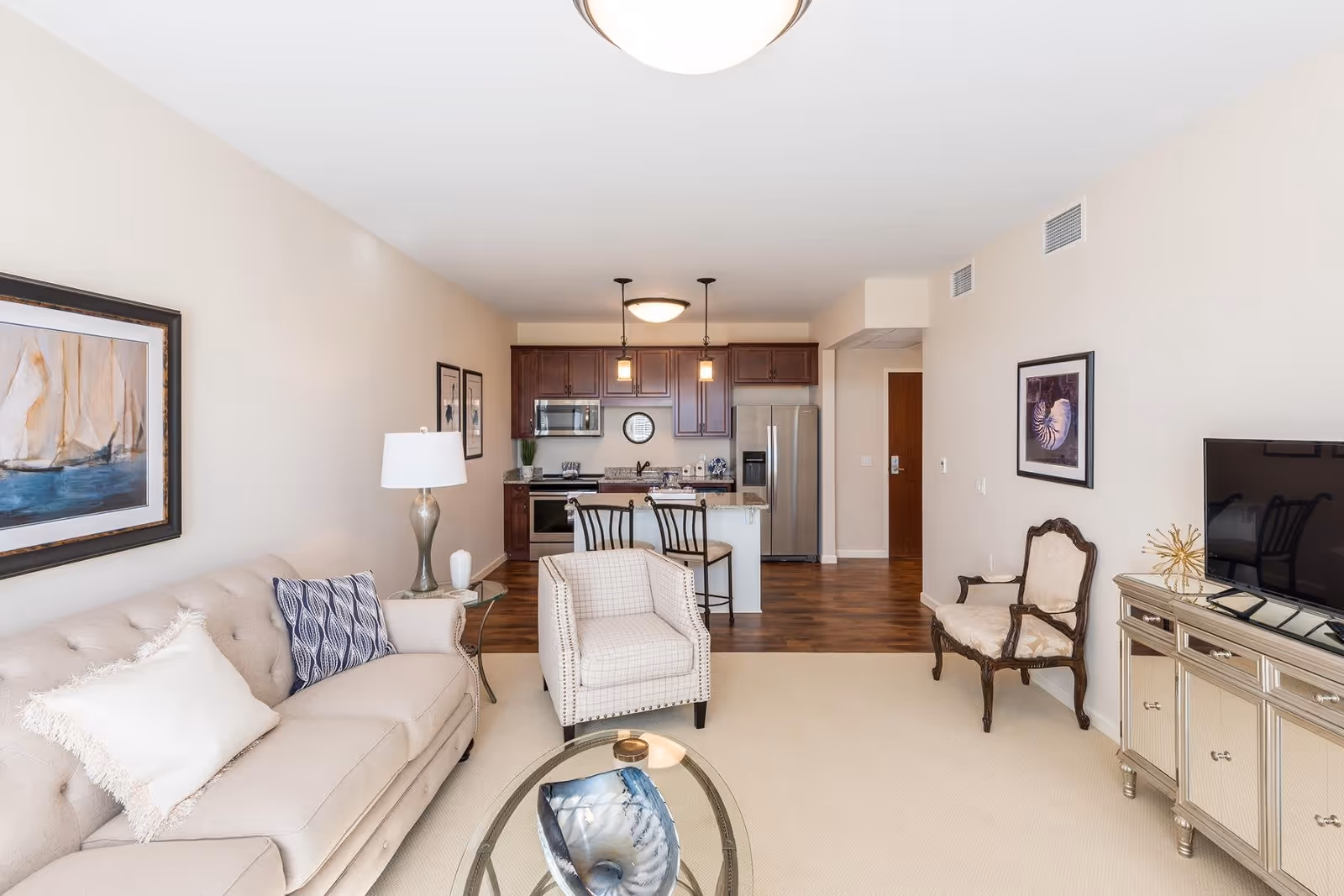 A bright and spacious living room with a beige sofa, a patterned armchair, and a wooden chair with upholstery. There is a glass coffee table with a decorative item on it. The room has light-colored walls adorned with framed artwork. In the background, there is an open kitchen with dark wood cabinets, stainless steel appliances, and a small island with two bar stools. The floor transitions from carpet in the living area to wood in the kitchen.