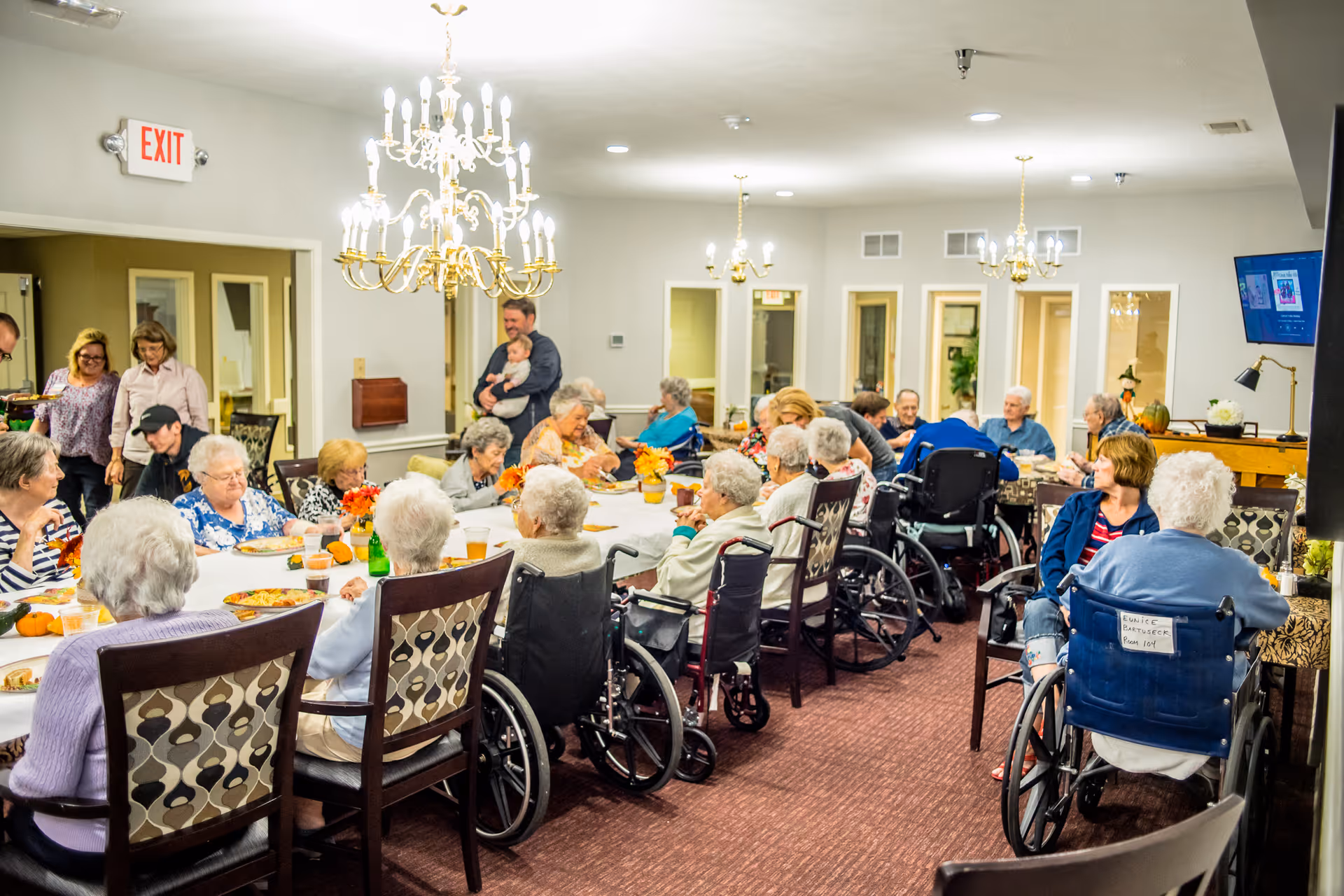 A group of elderly people, many in wheelchairs, seated around a long dining table in a well-lit room with chandeliers. Several caregivers and visitors are standing and interacting with the residents. The room has a warm, communal atmosphere with decorations on the table and a television mounted on the wall.
