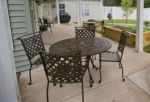 Outdoor covered patio area with a round metal table and four matching metal chairs. In the background, there is a grill, additional seating, raised garden beds with flowers, and a white fence enclosing the yard.