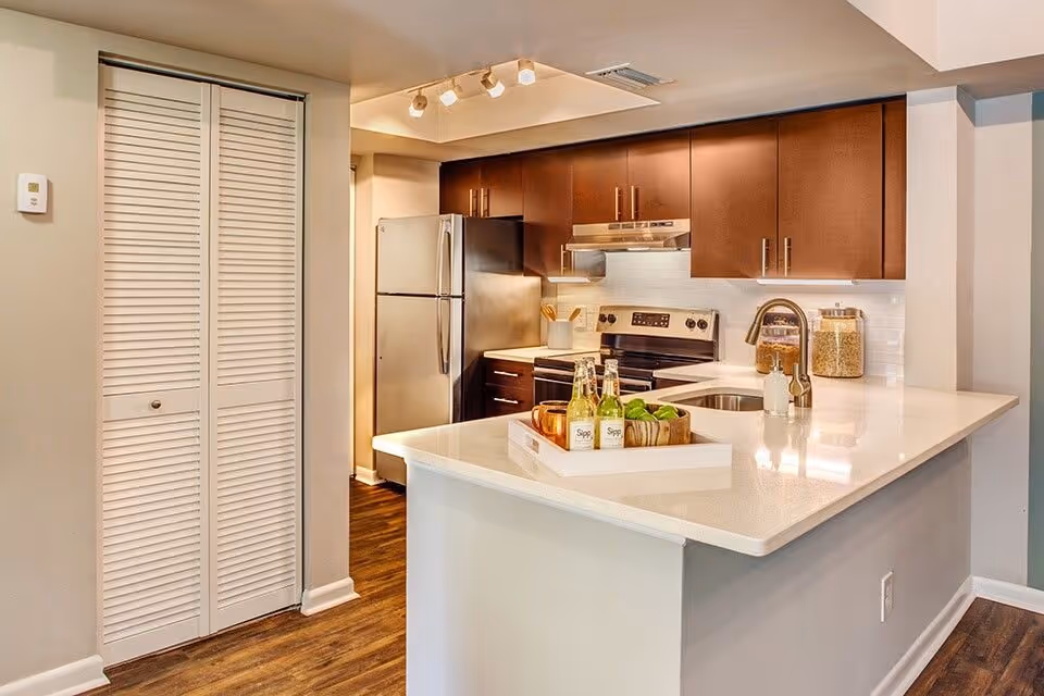 Modern kitchen with dark wood cabinets, stainless steel refrigerator and stove, white countertops, a sink with a faucet, and a tray with bottles and green apples on the counter. The floor is wooden and there is a white louvered door on the left side.