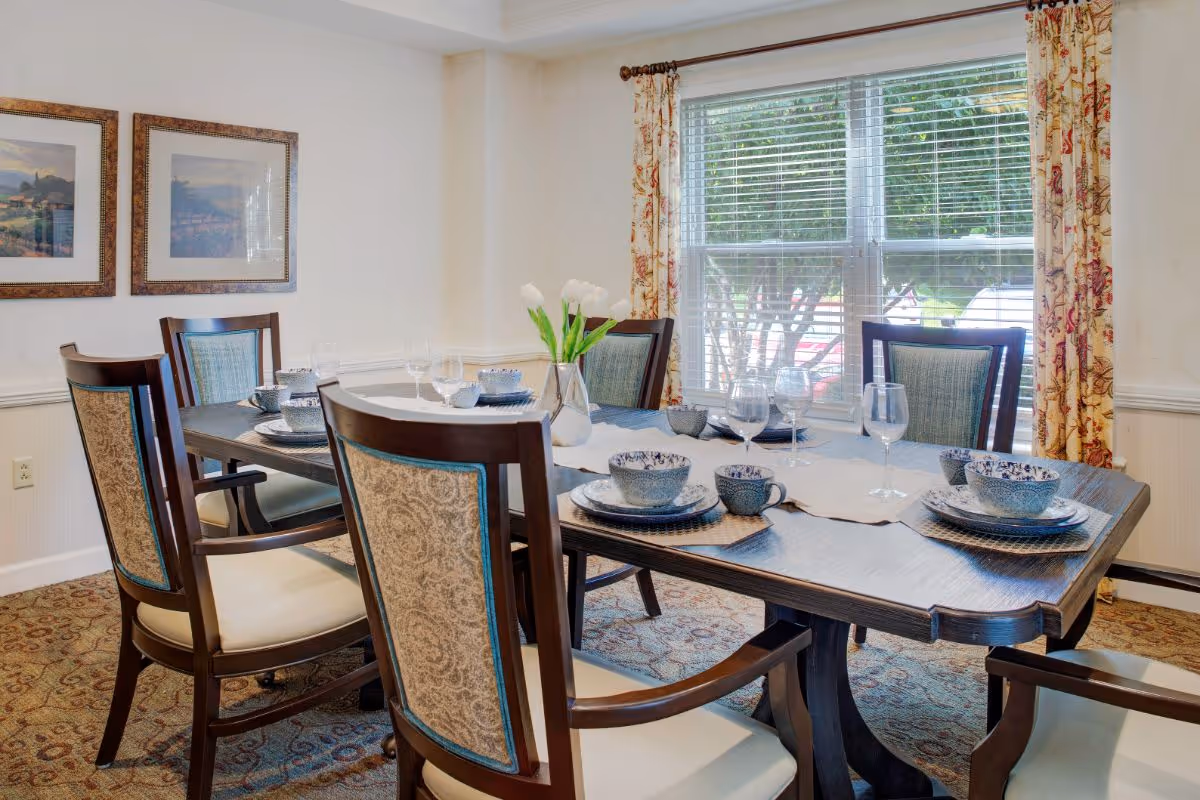 A dining room with a wooden table set for six people, featuring blue and white patterned dishes, glassware, and a vase with white tulips in the center. The room has four wooden chairs with cushioned seats and backs, a window with blinds and floral curtains, and two framed landscape paintings on the wall.