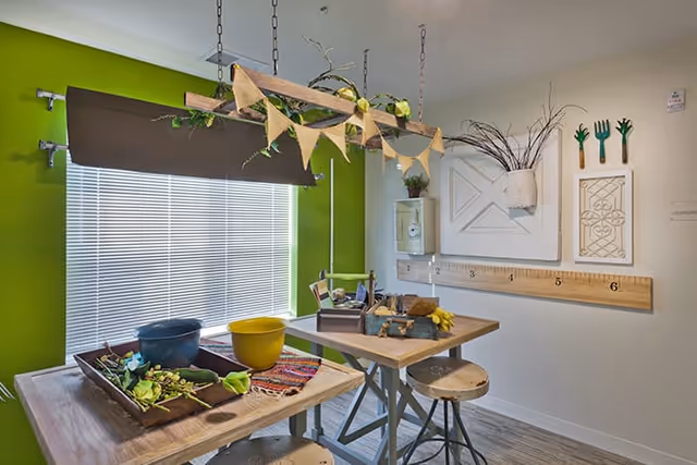A bright craft or activity room with two wooden tables and stools. The tables have various craft supplies including pots, plants, and gardening tools. The walls are decorated with a large wooden ruler, a white decorative panel, and hanging planters. A green accent wall is visible behind a window with closed blinds.