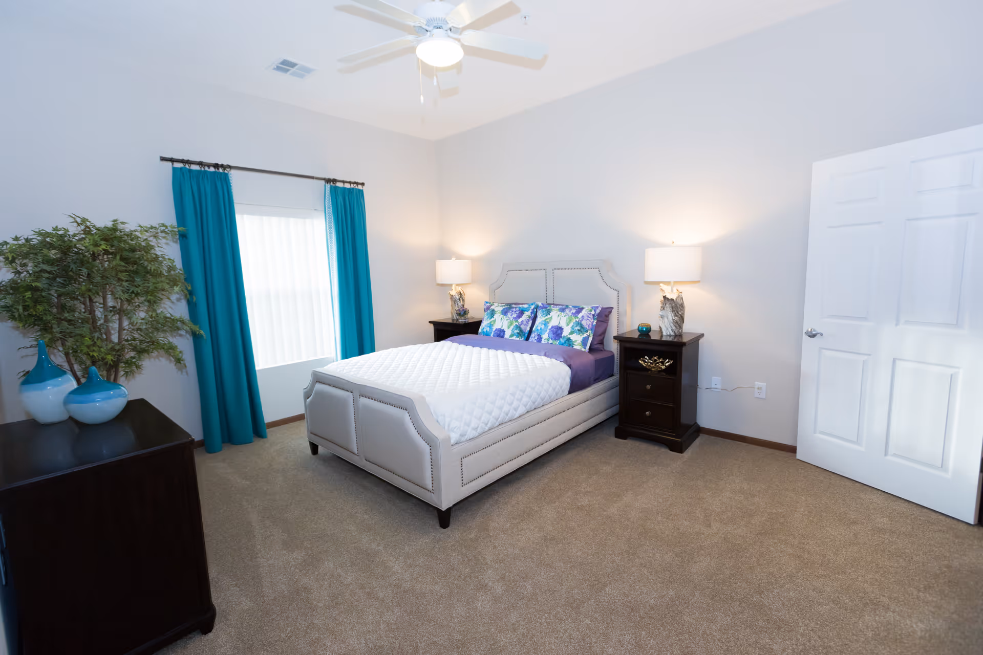 A bright and clean bedroom with a beige upholstered bed featuring a white quilt and floral pillows. There are two dark wooden nightstands on either side of the bed, each with a lamp. A window with teal curtains is on the left wall, and a ceiling fan with a light is mounted on the ceiling. A dark wooden dresser with decorative vases and a green plant is positioned near the window. The room has beige carpet and light-colored walls.