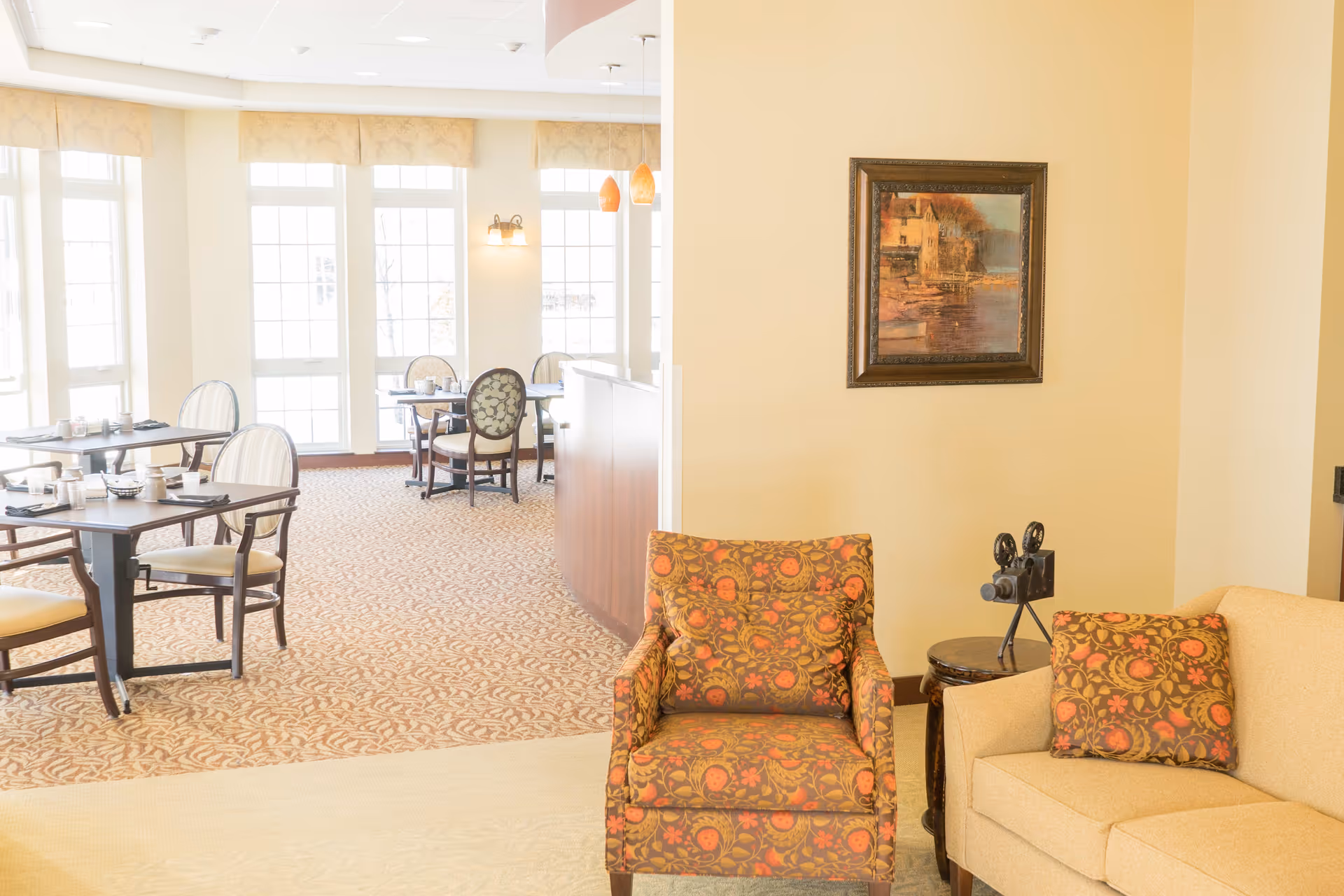 A bright and inviting senior living facility interior showing a dining area with tables and chairs set for meals, adjacent to a cozy seating area with a patterned armchair, a beige sofa with a matching patterned pillow, a small side table with a vintage film camera, and a framed painting on the wall.