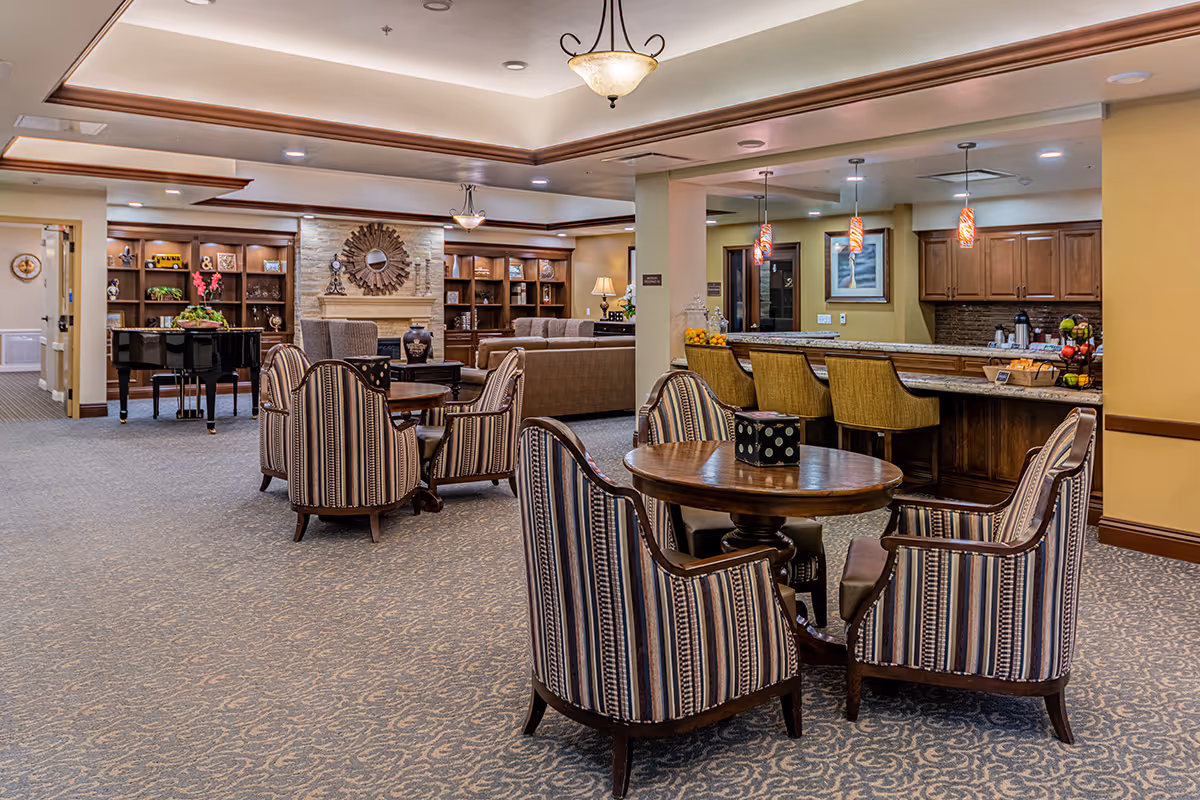 A spacious senior living common area with patterned carpet, several round wooden tables surrounded by striped upholstered chairs, a bar counter with high chairs, pendant lights, a stone fireplace with a decorative mirror above it, built-in wooden bookshelves, and a black grand piano in the background.