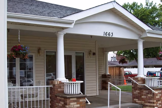 Front entrance of a building with white columns and a covered porch. The building has the number 1663 displayed above the entrance. There are hanging flower baskets and a white metal railing along the walkway leading to the door. Several parked vehicles and trees are visible in the background.
