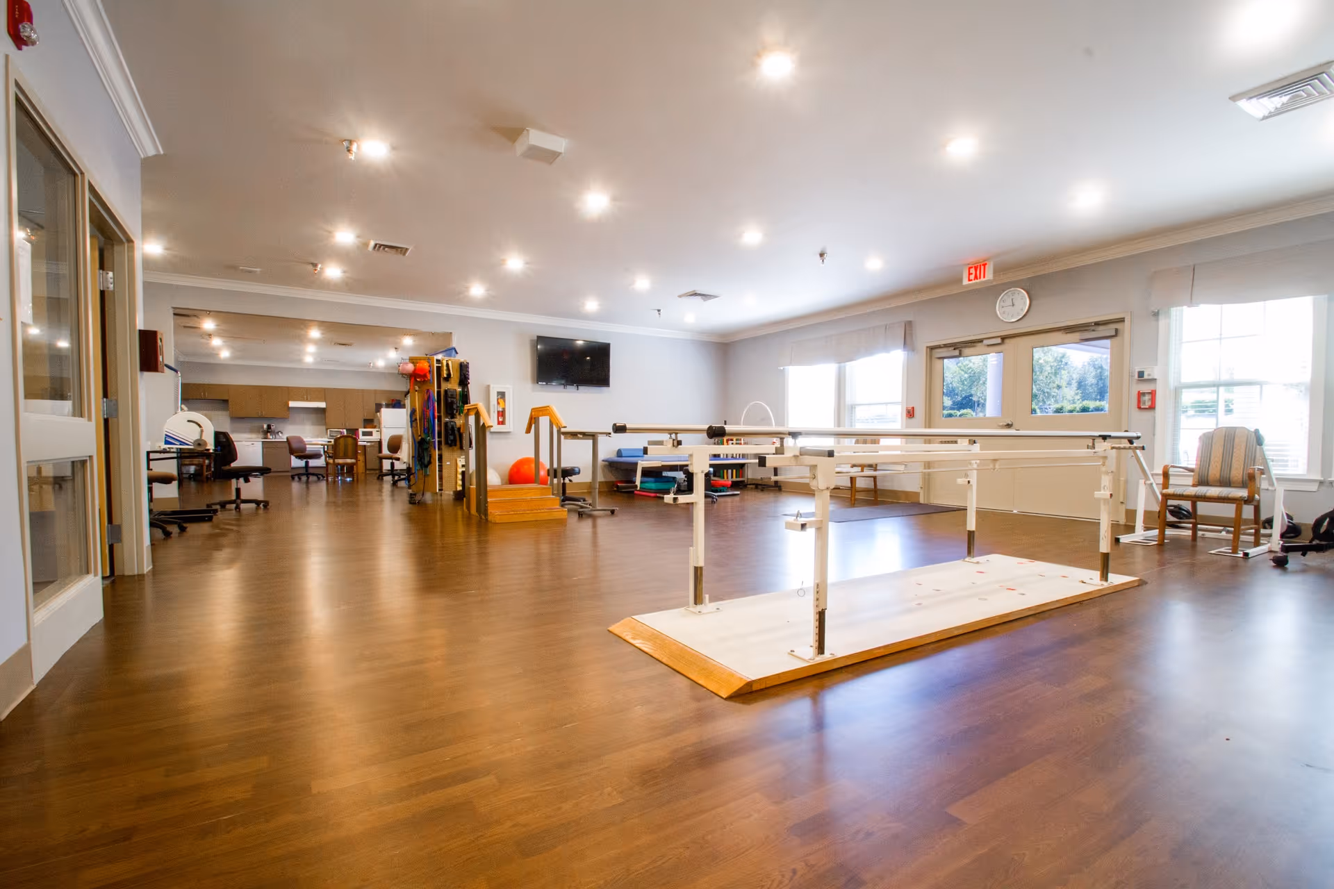 A spacious rehabilitation room with wooden flooring and bright overhead lighting. The room contains parallel bars for physical therapy, exercise equipment, chairs, and a wall-mounted TV. Large windows allow natural light to enter, and there is a clock above the exit door.