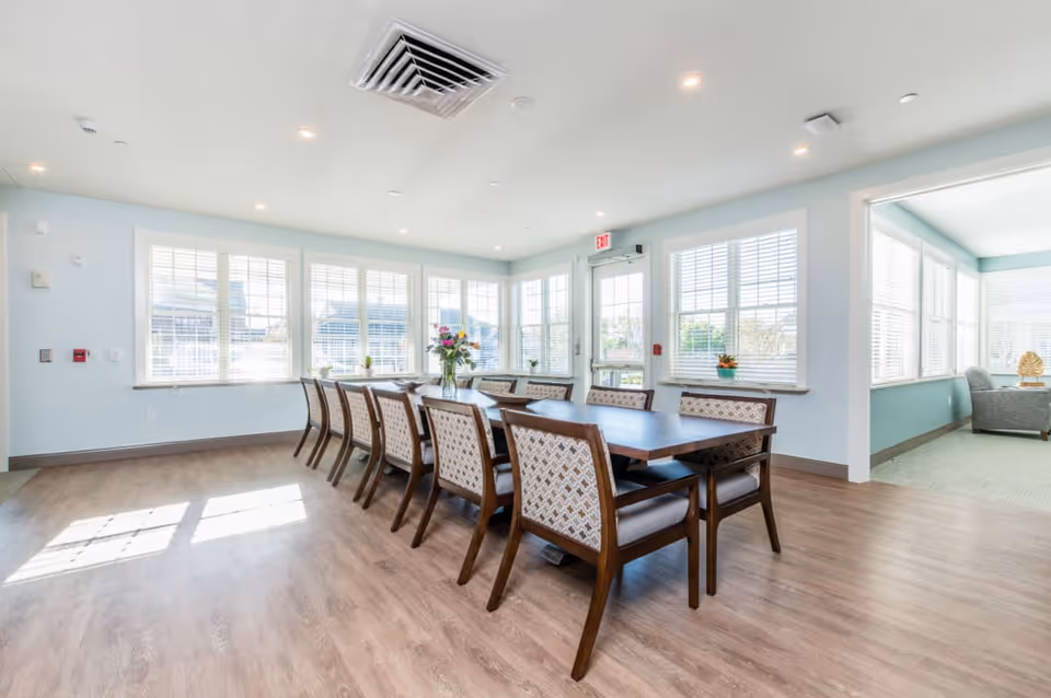 Bright communal dining room with a long wooden table, upholstered chairs, and large windows letting in natural light.