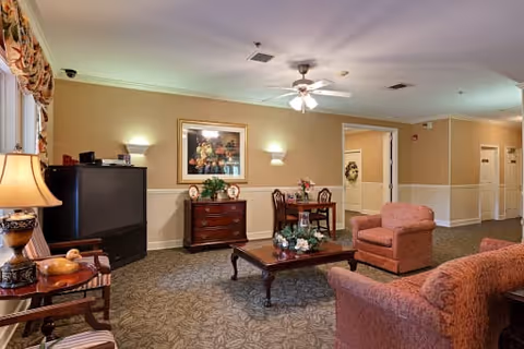A cozy living room area in an assisted living facility featuring two upholstered armchairs and a matching sofa, a wooden coffee table with floral decorations, a small dining table with two chairs, a wooden chest of drawers with a framed painting above it, a large TV, and warm lighting from lamps and wall sconces.