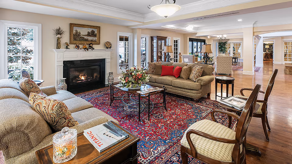 A spacious and well-lit living room in a senior living facility featuring two beige sofas with patterned cushions, a red and blue patterned area rug, a glass coffee table with a flower arrangement, and a fireplace with a white mantel decorated with small figurines and a framed painting above it. The room has large windows, wooden floors, and additional seating with wooden chairs and side tables.