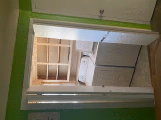 Laundry area with a white washing machine and dryer side by side, wooden shelves above them, and green walls surrounding the space.