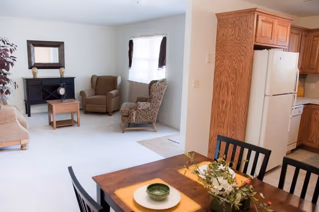 A bright living and dining area with beige carpet and white walls. The living area has a beige sofa, two armchairs, a black console table with a mirror above it, and a small wooden coffee table. The dining area features a wooden table set with placemats, a green bowl, and a floral centerpiece. The kitchen is visible with wooden cabinets, a white refrigerator, and a dishwasher.