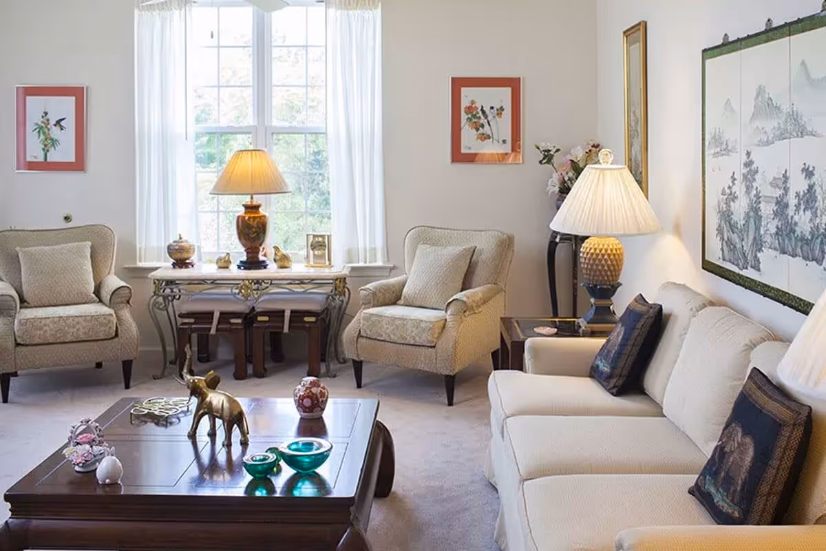 A bright and cozy living room with a large window covered by sheer white curtains. The room features a beige sofa with decorative pillows, two matching armchairs, a wooden coffee table with decorative items including a small elephant figurine, and two side tables with lamps. The walls are adorned with framed artwork and a large decorative panel.