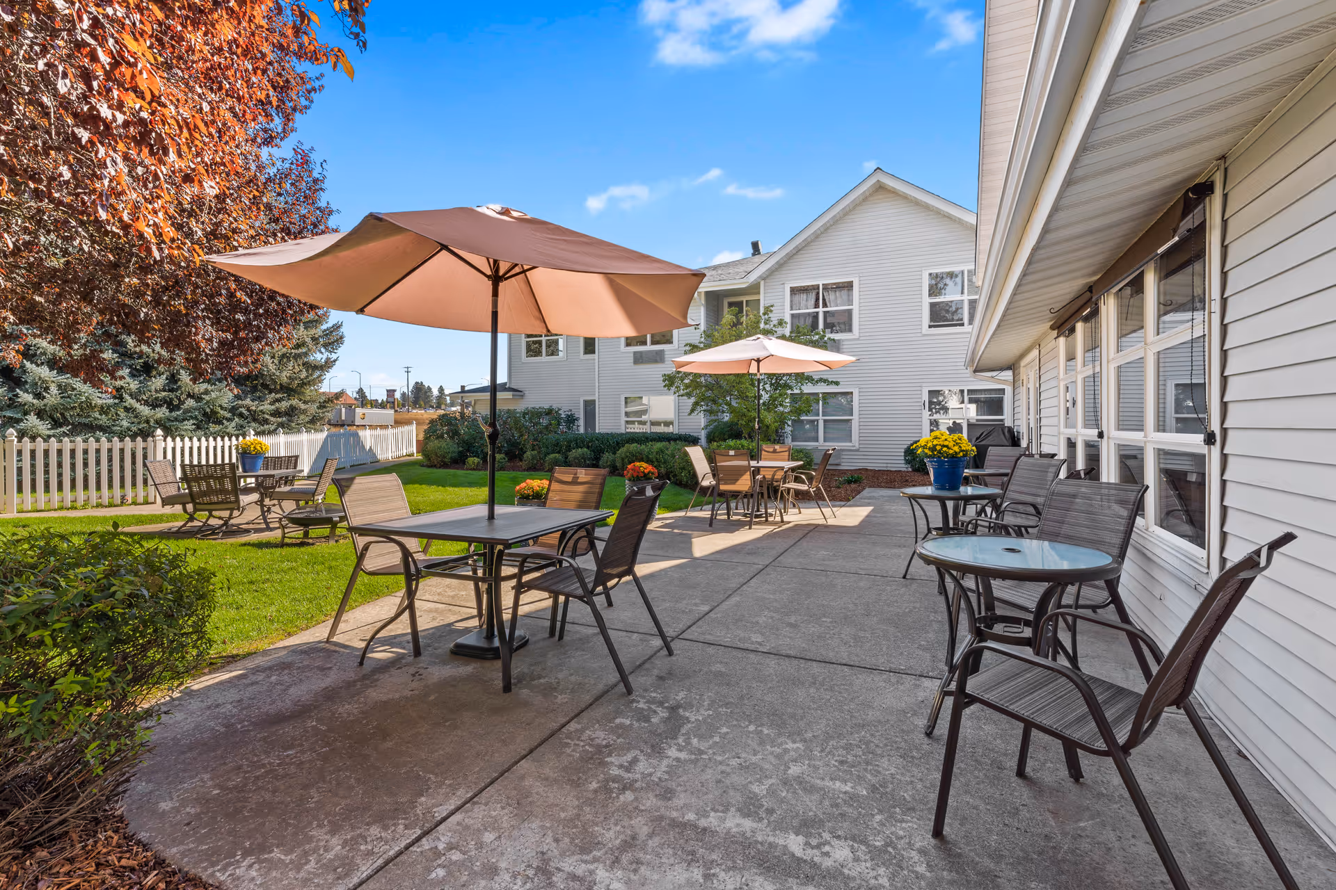 Outdoor patio area at Legends Park Assisted Living with several tables and chairs, some shaded by large umbrellas. The patio is adjacent to a white building with multiple windows. There is a green lawn with bushes and trees, including one with red leaves, and a white picket fence in the background under a clear blue sky.