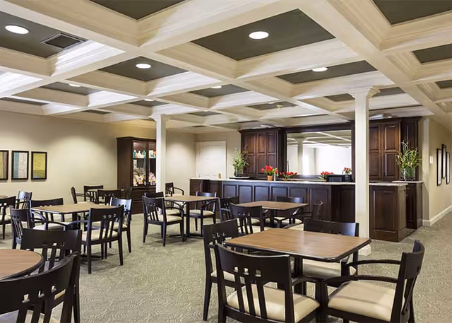 A spacious dining area with multiple wooden tables and chairs arranged neatly. The room features a coffered ceiling with recessed lighting, beige walls, and carpeted floors. There is a dark wood buffet or serving counter along one wall with decorative plants and flowers on top. Framed artwork is hung on the walls, and a glass-front cabinet is visible in the background.