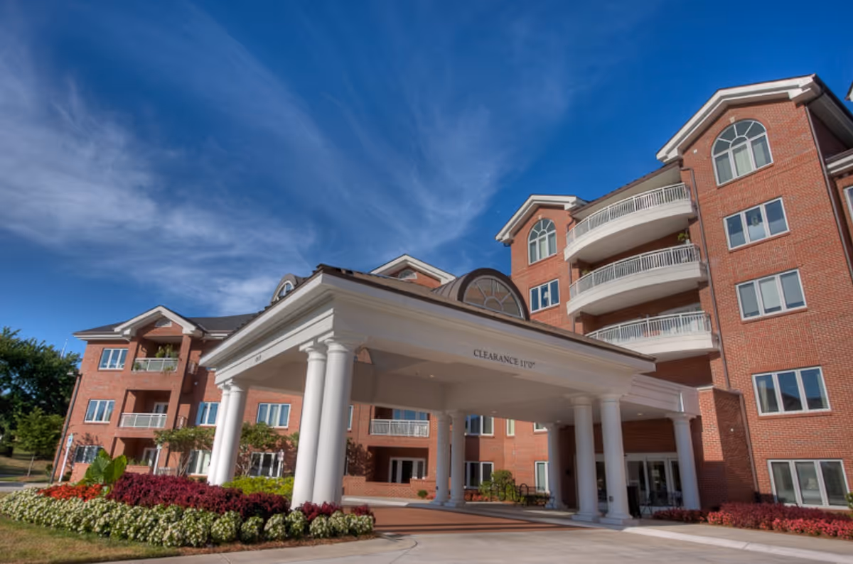 Front entrance of a multi-story red-brick building with a white columned porte-cochère, balconies, and landscaped flower beds under a blue sky.