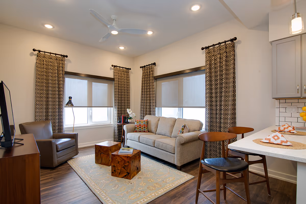 Cozy open living room with a sofa, armchair, patterned curtains, wooden coffee tables on a rug, and a kitchen counter with stools.