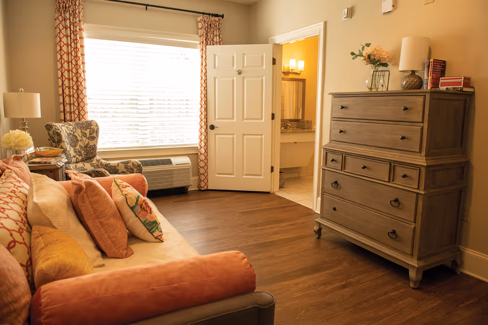 A cozy living room area with a sofa adorned with multiple colorful pillows, a floral patterned armchair next to a window with red and white patterned curtains, a wooden dresser with decorative items on top, and an open door leading to a bathroom with a lit vanity mirror.