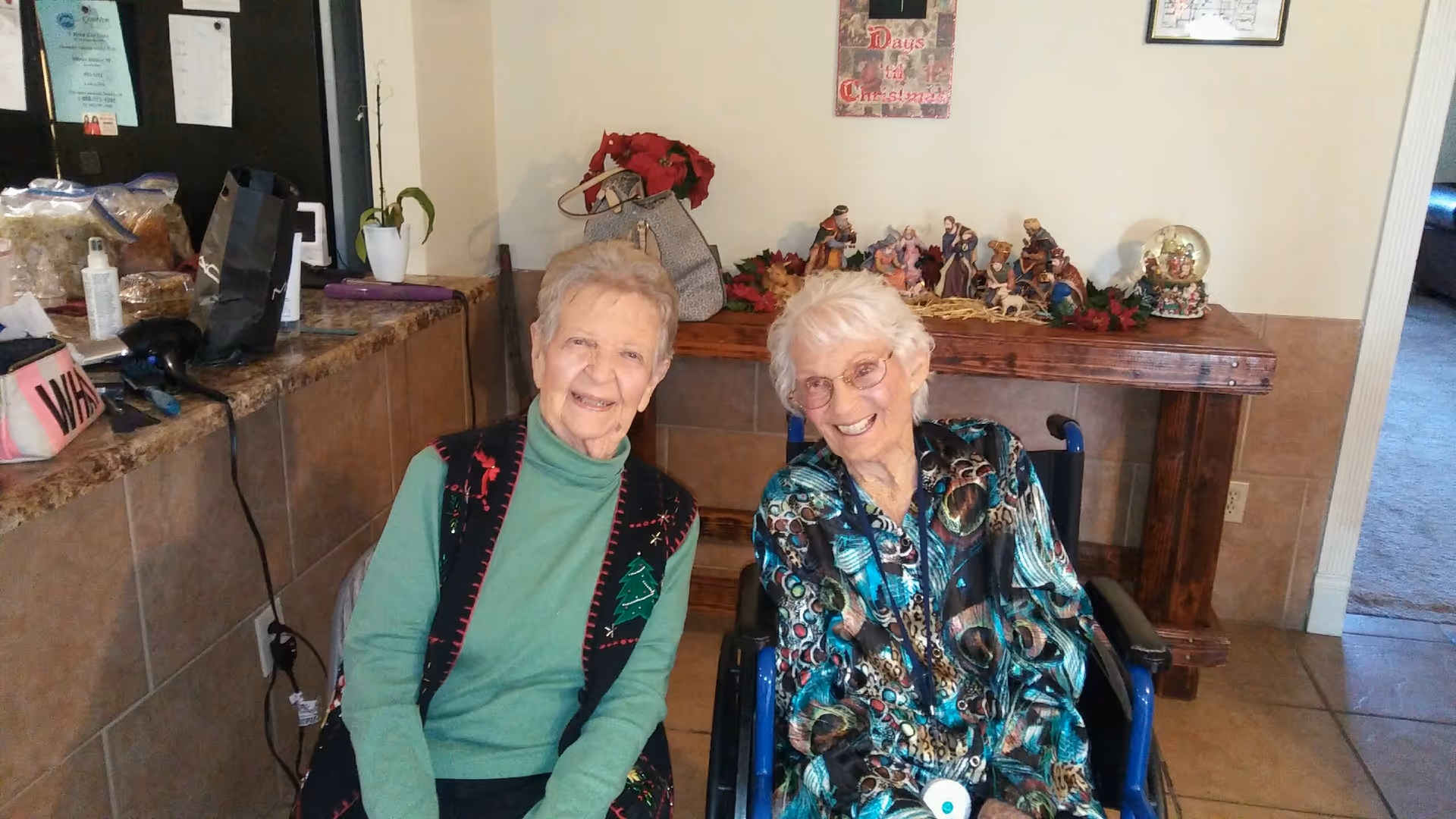 Two elderly women smiling and sitting indoors. One woman is seated in a wheelchair wearing a colorful patterned blouse, and the other woman is sitting next to her wearing a green turtleneck and a black vest with holiday-themed decorations. Behind them is a wooden table with Christmas decorations and a sign on the wall that reads 'Days till Christmas.'