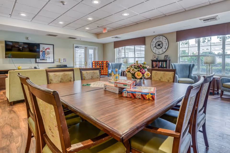 A well-lit common area with a large wooden table surrounded by chairs. On the table are several board games and a vase with colorful flowers. In the background, there are comfortable armchairs, a bookshelf, a large wall clock, and a flat-screen TV mounted on the wall. Large windows allow natural light to fill the room.