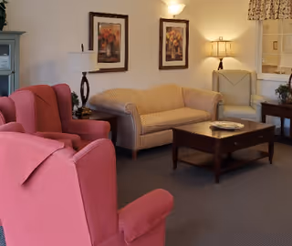 A cozy living room with two red armchairs, a beige striped sofa, a wooden coffee table with a decorative plate, two framed floral paintings on the wall, a floor lamp, and a side table with a lamp and a plant.