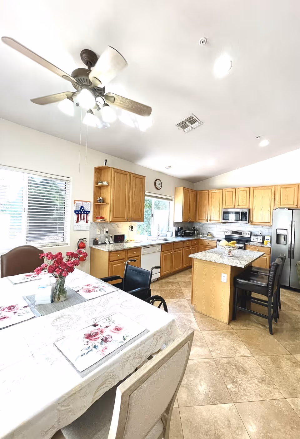Bright open kitchen with wooden cabinets, a center island, stainless steel appliances and a dining table with a floral tablecloth and vase of flowers.
