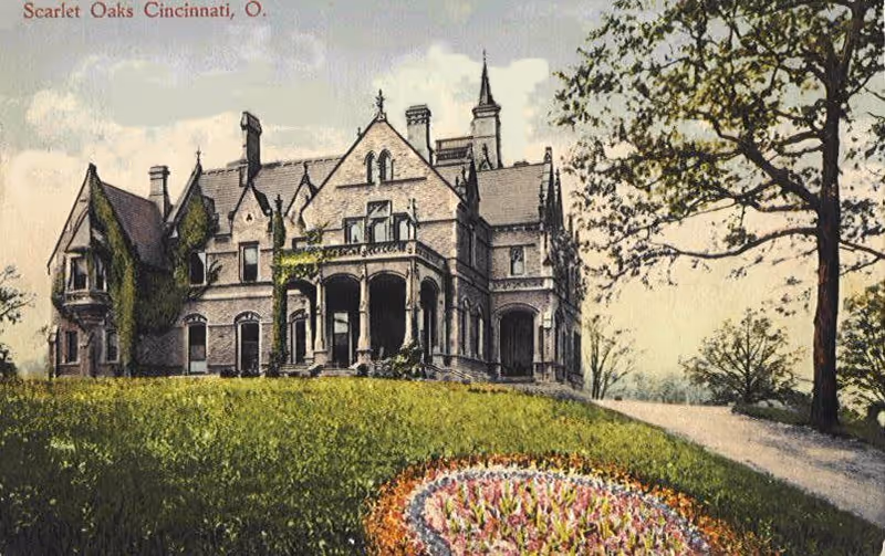 A large, historic mansion-style building with multiple chimneys and a pointed tower, surrounded by a grassy lawn with a circular flower bed in the foreground and trees on the right side.