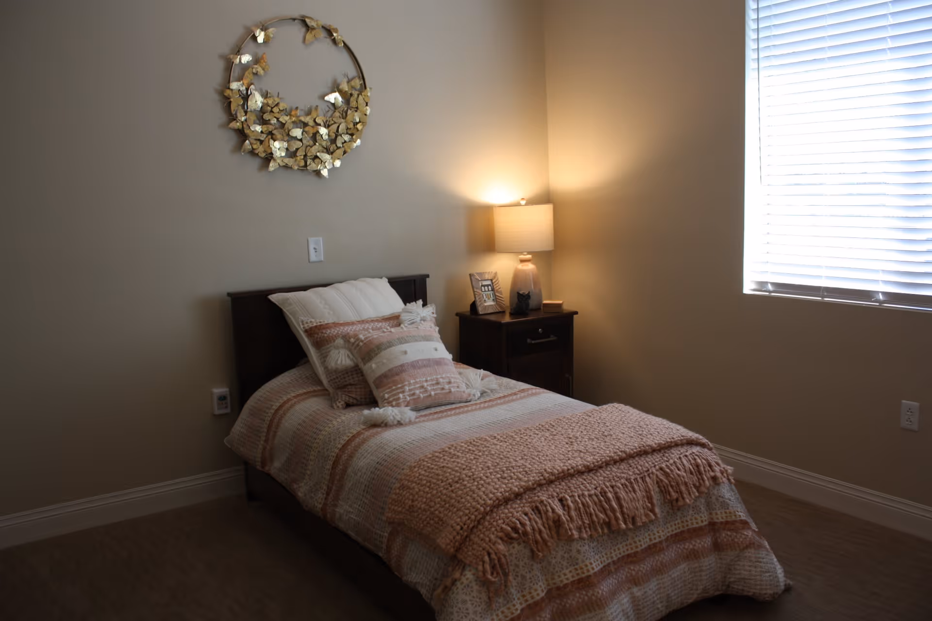 A cozy bedroom with a single bed covered in a patterned quilt and multiple pillows. A wooden nightstand with a lamp, a framed photo, and a small decorative item sits beside the bed. A window with blinds allows natural light into the room, and a decorative wreath hangs on the wall above the bed.