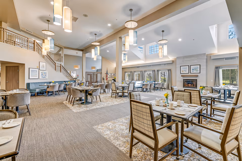 A spacious and well-lit dining room in a senior living facility with multiple tables and chairs arranged neatly. The room features high ceilings with modern pendant lights, large windows with curtains allowing natural light, a fireplace with framed artwork above it, and a staircase leading to an upper level. The tables are set with plates, cups, and yellow flower centerpieces.