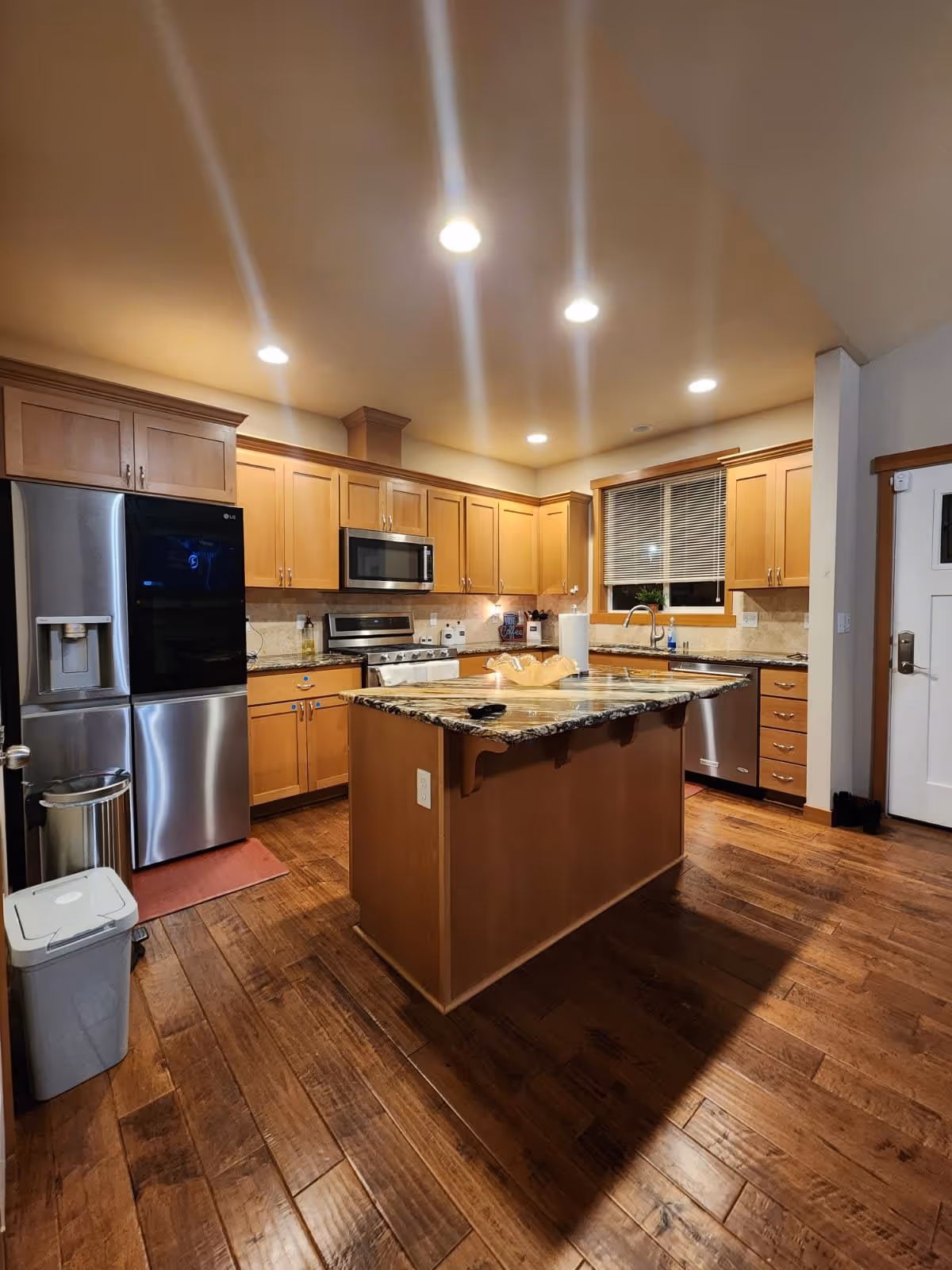 A modern kitchen with wooden cabinets, a large kitchen island with a granite countertop, stainless steel refrigerator, microwave, stove, and dishwasher. The kitchen has wooden flooring and recessed ceiling lights. A window with blinds is above the sink.
