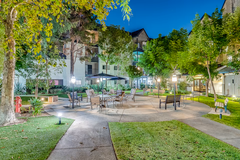 Well-lit courtyard of a senior living facility with outdoor seating, tables and umbrellas surrounded by apartment buildings and trees at dusk.