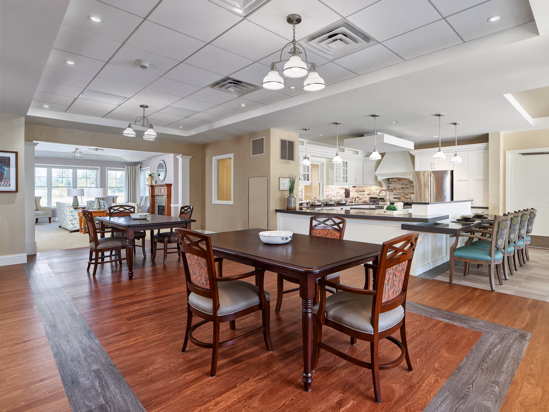 A spacious dining area with wooden tables and chairs, adjacent to a modern kitchen with white cabinetry, stainless steel appliances, and pendant lighting. In the background, there is a cozy living room area with large windows, armchairs, and a fireplace.
