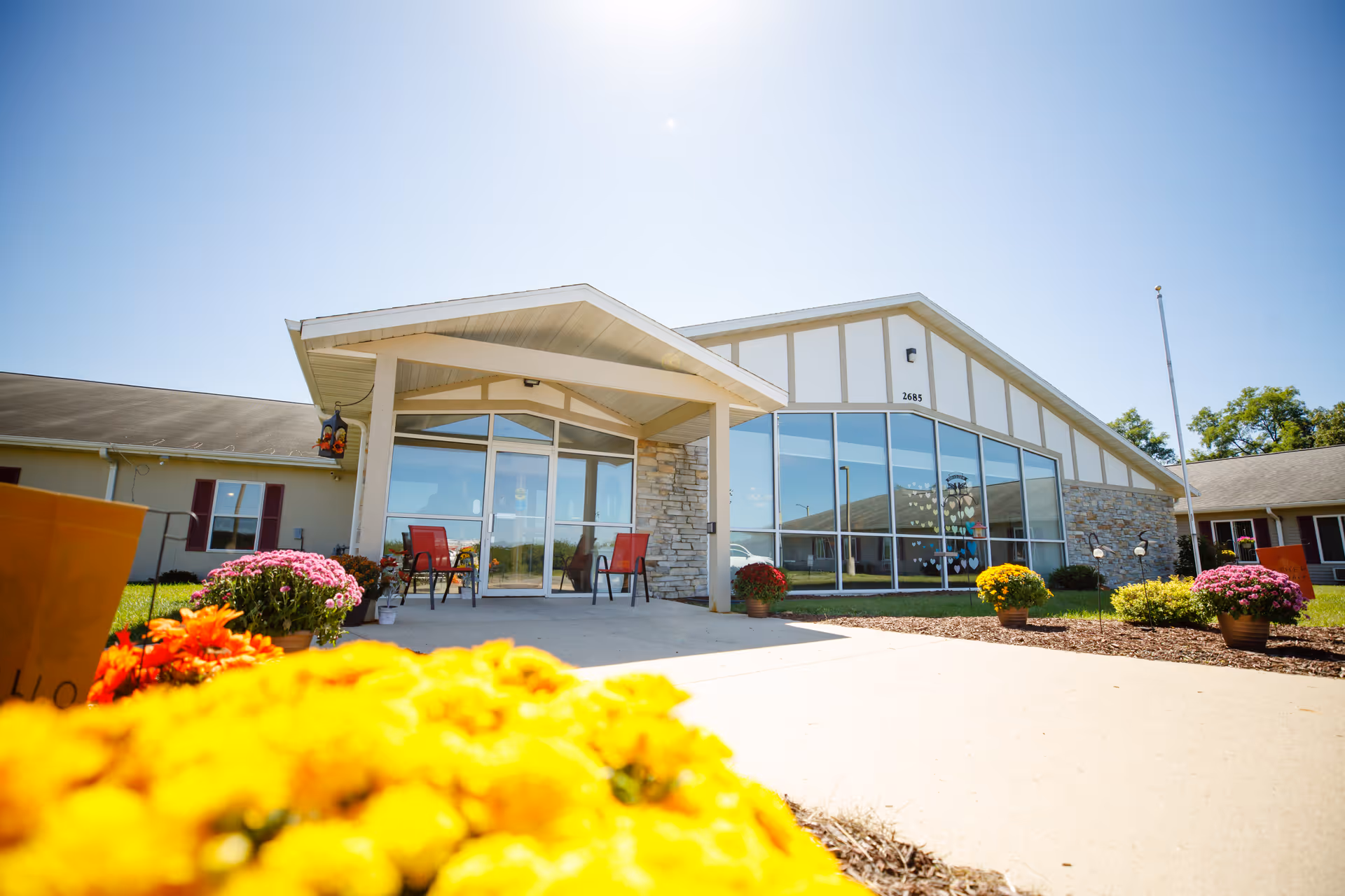 The front exterior of a modern assisted living facility with a glass entry and colorful potted flowers along the walkway.
