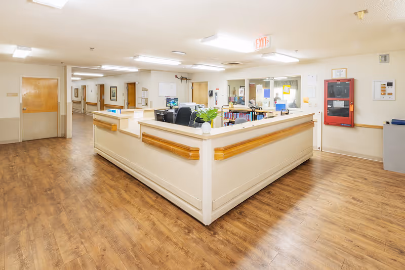 Central nurses' station in a well-lit senior living facility hallway with wood floors and surrounding doors.